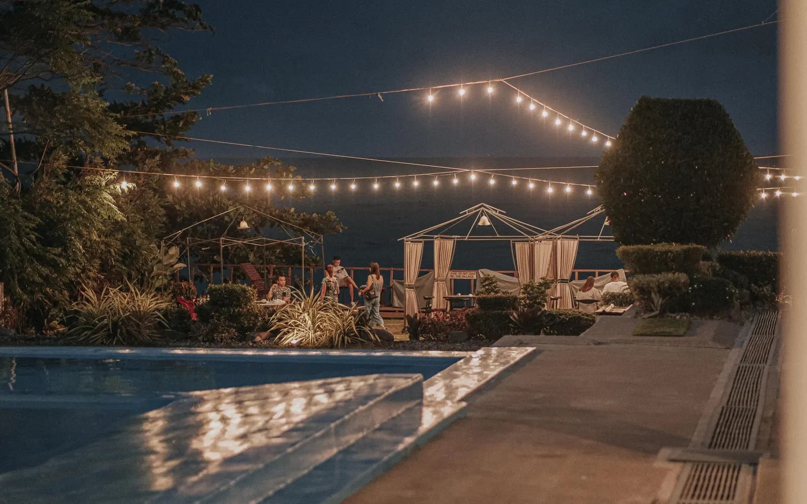 Temporary concept image of friends gathered beside a pool in the evening under string lights; not property photography.