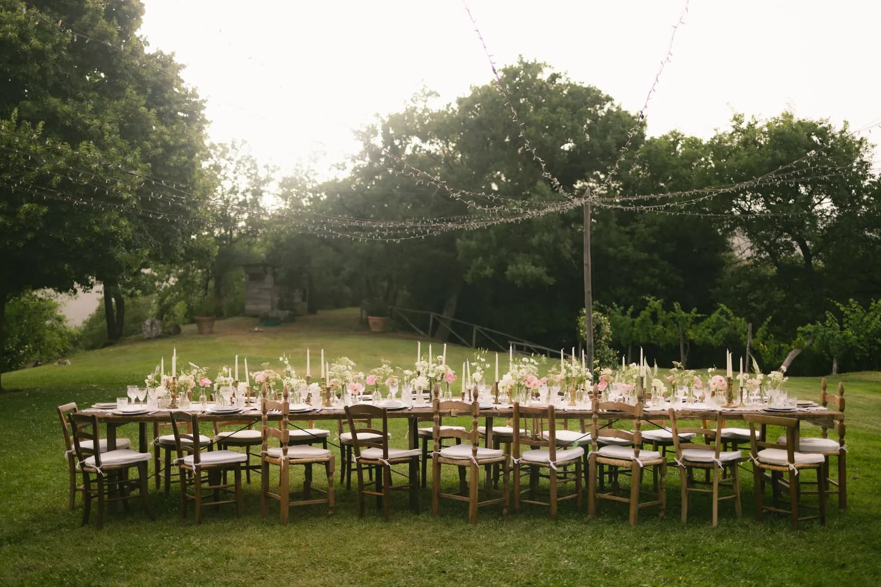 Temporary concept image of long dinner table set on a lawn beneath strings of warm festoon lights; not property photography.