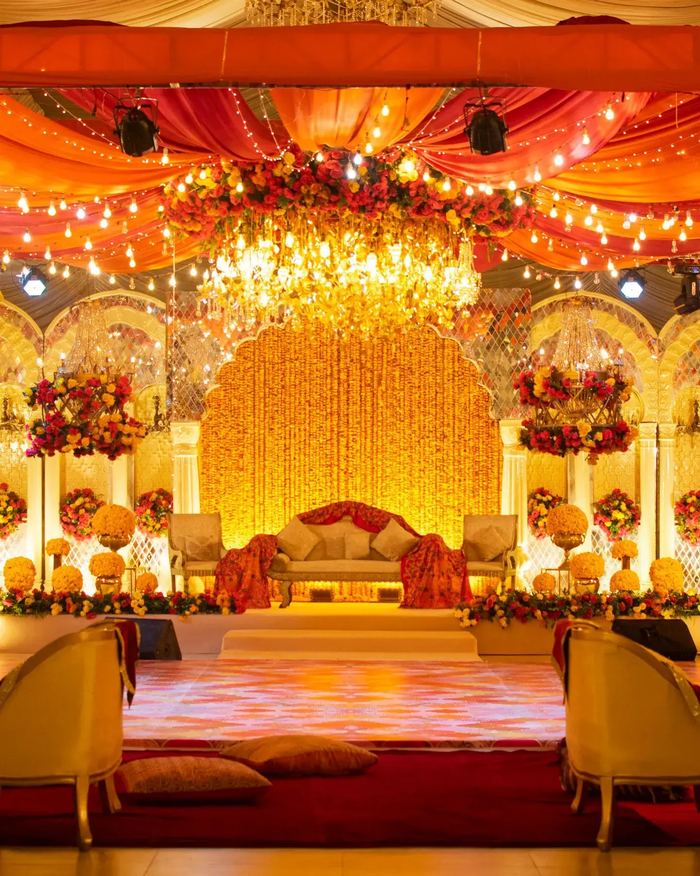 Temporary concept image of grand marriage-hall stage with a marigold backdrop, chandelier and saffron drapes glowing in warm light; not property photography.
