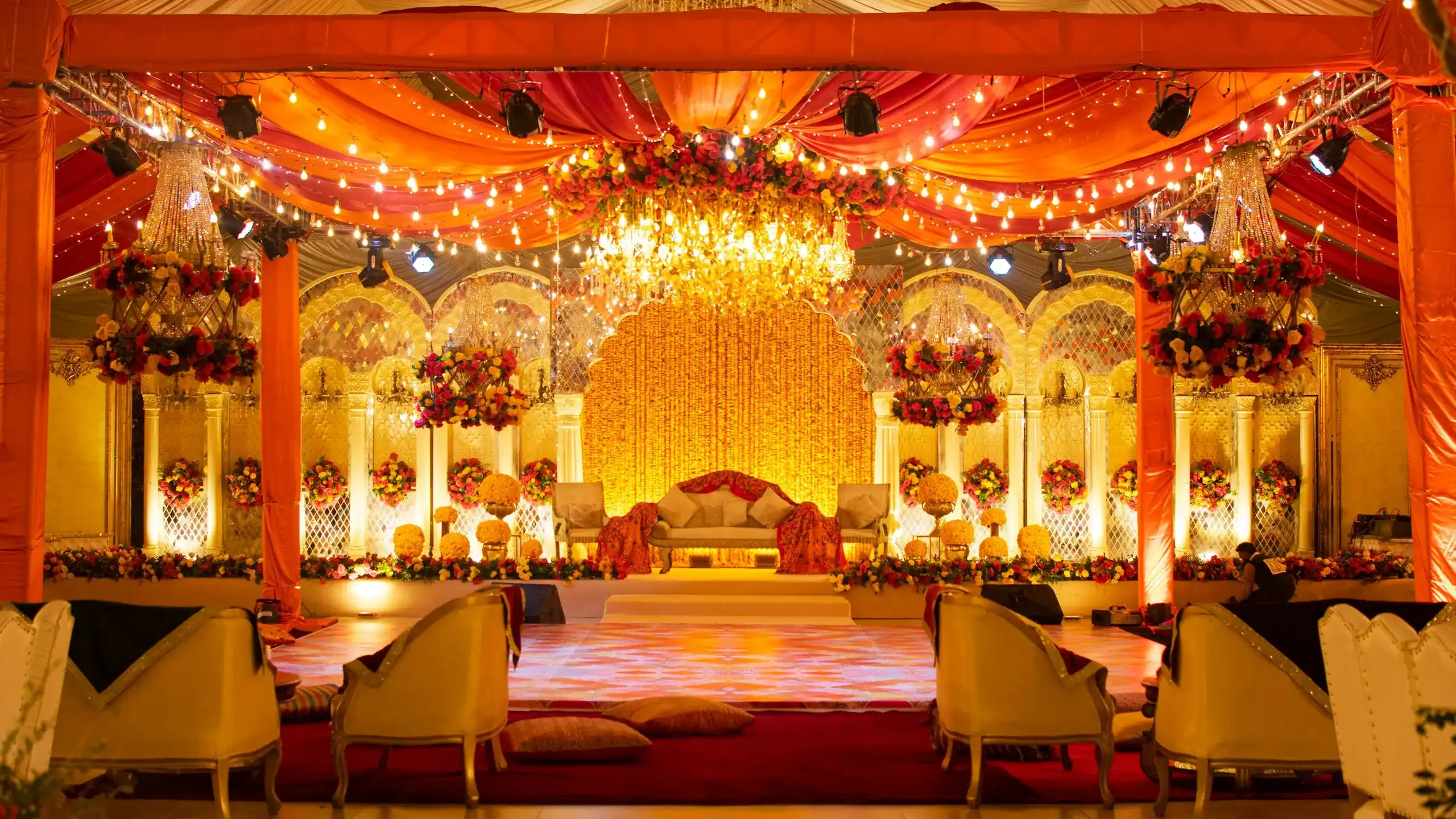 Temporary concept image of grand marriage-hall stage with a marigold backdrop, chandelier and saffron drapes glowing in warm light; not property photography.