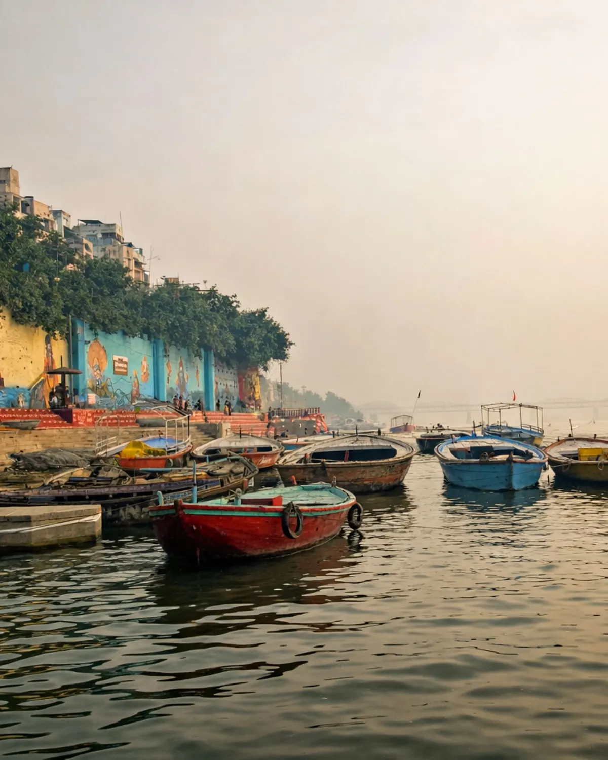 Temporary concept image of wooden boats moored on the Ganga at dawn with ghats rising behind; not property photography.