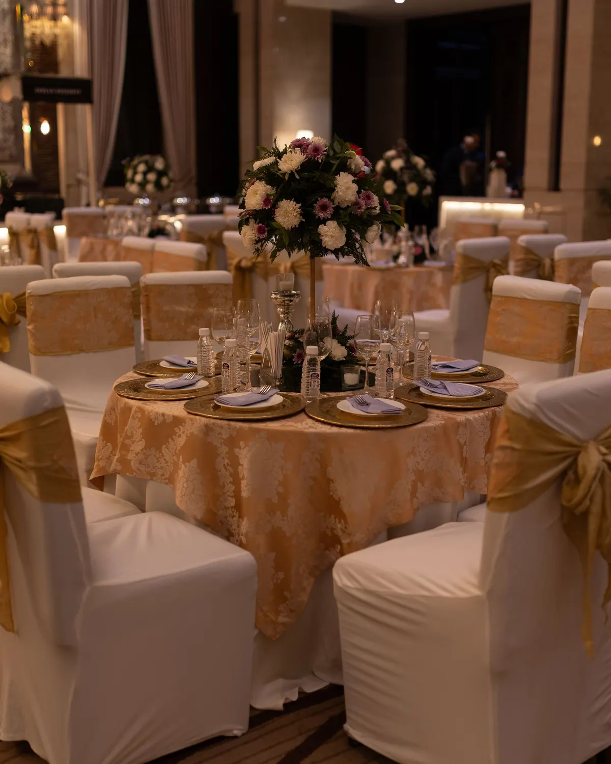 Temporary concept image of banquet tables dressed in ivory and gold with floral centrepieces under warm light; not property photography.