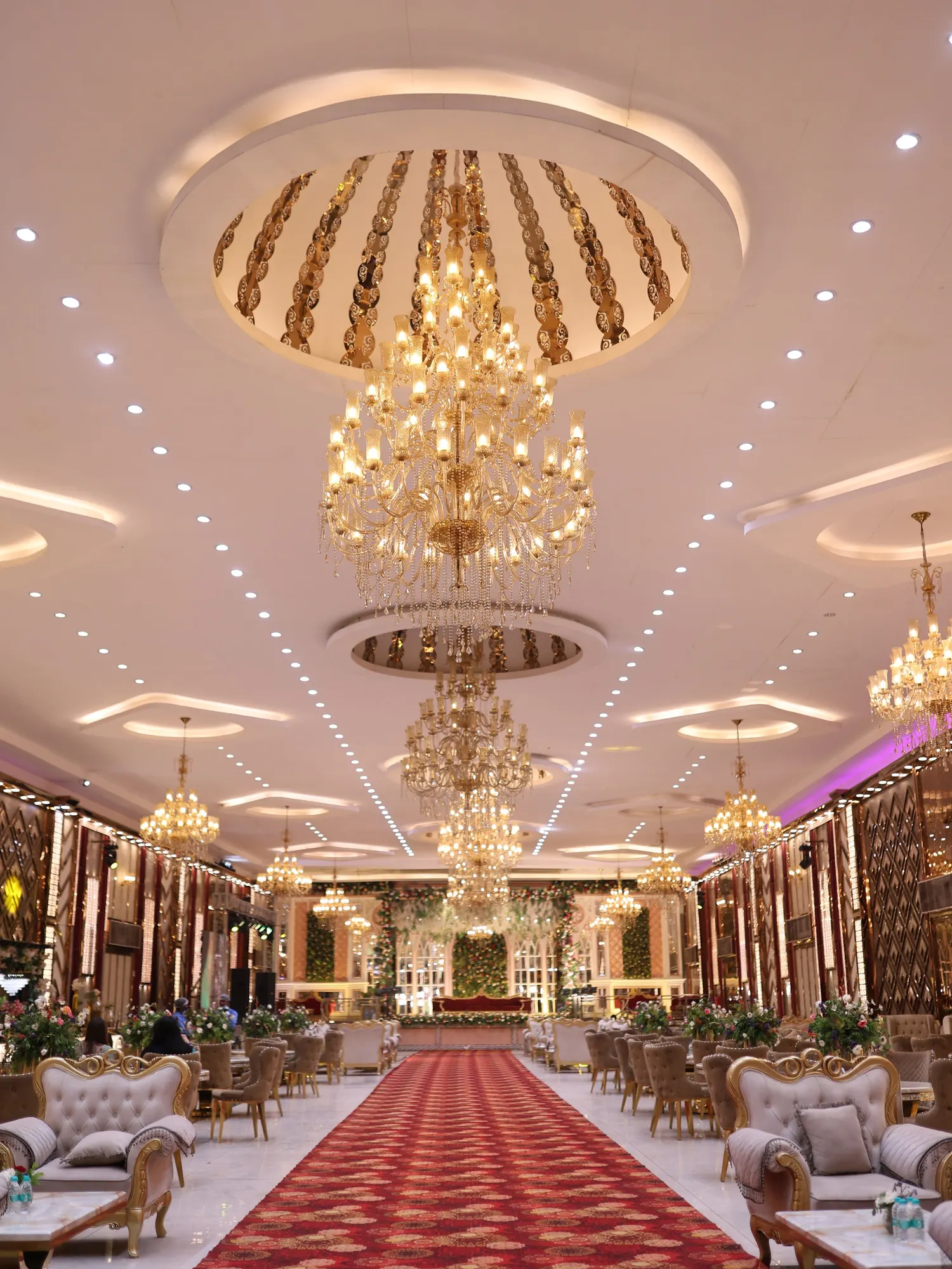 Temporary concept image of grand banquet hall with tiered gold chandeliers, a red carpet aisle and cream seating; not property photography.